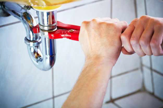 plumber holding pair of pliers with both hands loosening a drain pipe underneath a sink.
