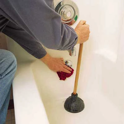 Home owner with wash cloth placed on drain stop of bath tub with left hand and a plunger in his right hand placed on the drain.